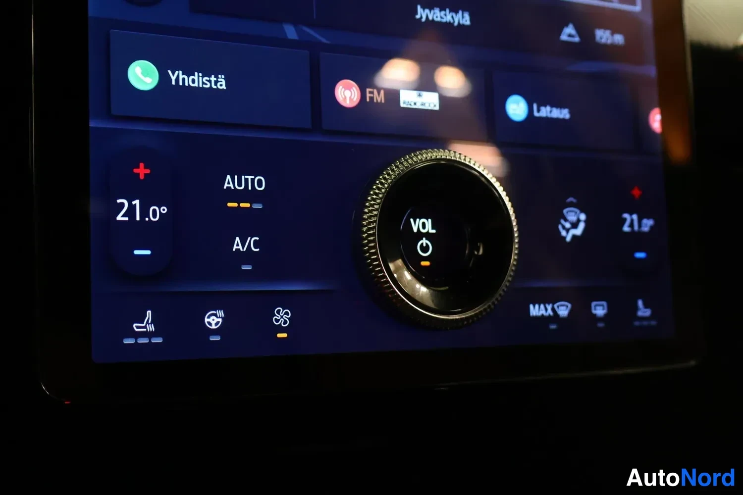 Edestä: Yksityiskohta infotainment-näytön ilmastointiosiosta: lämpötila 21,0°, AUTO ja A/C valittuna, äänenvoimakkuussäädin VOL ja painike Lataus näkyvissä.