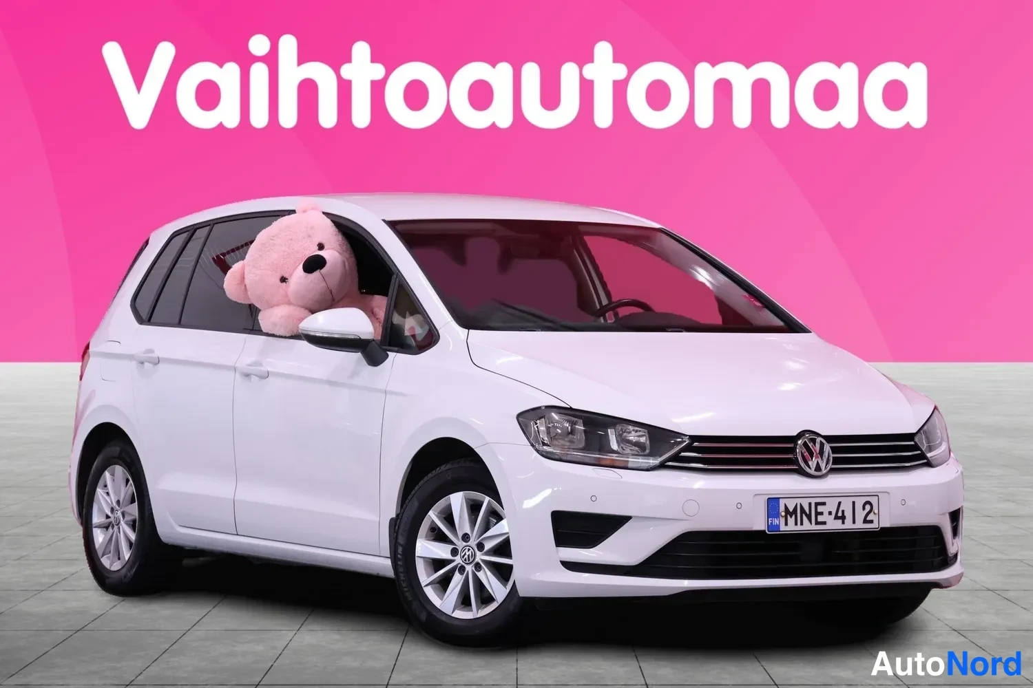Etuviisto, nokka oikealle: Valkoinen Volkswagen Golf Sportsvan etuviistossa, pehmolelu kuljettajan ikkunassa, rekisteri.