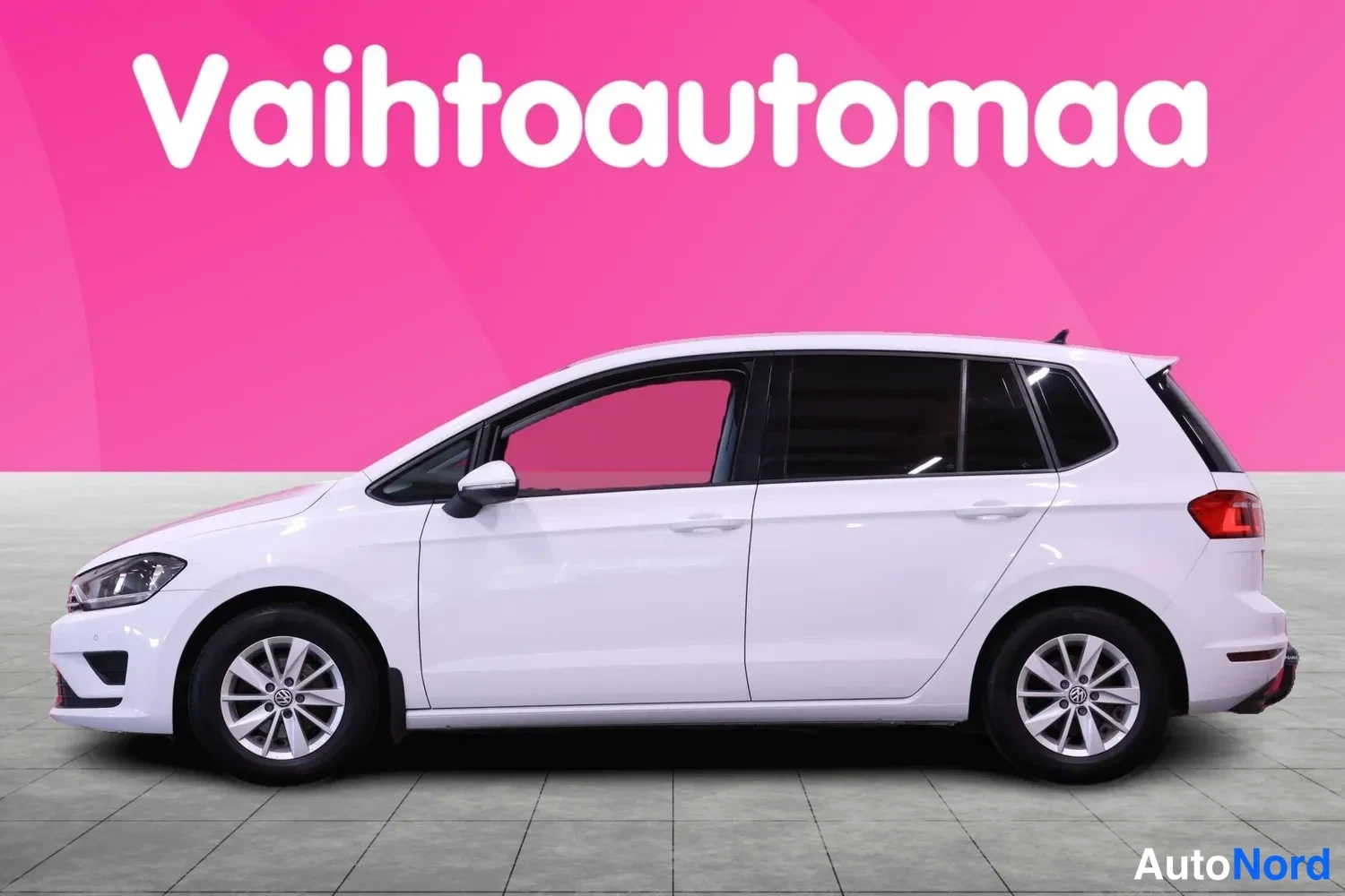 Etuviisto, nokka vasemmalle: Valkoisen tila-automaisen henkilöauton vasen kylki kulmasta kuvattuna; ajovalo näkyy keulassa ja takavalo takana, katolaatta-kanava pohjana.
