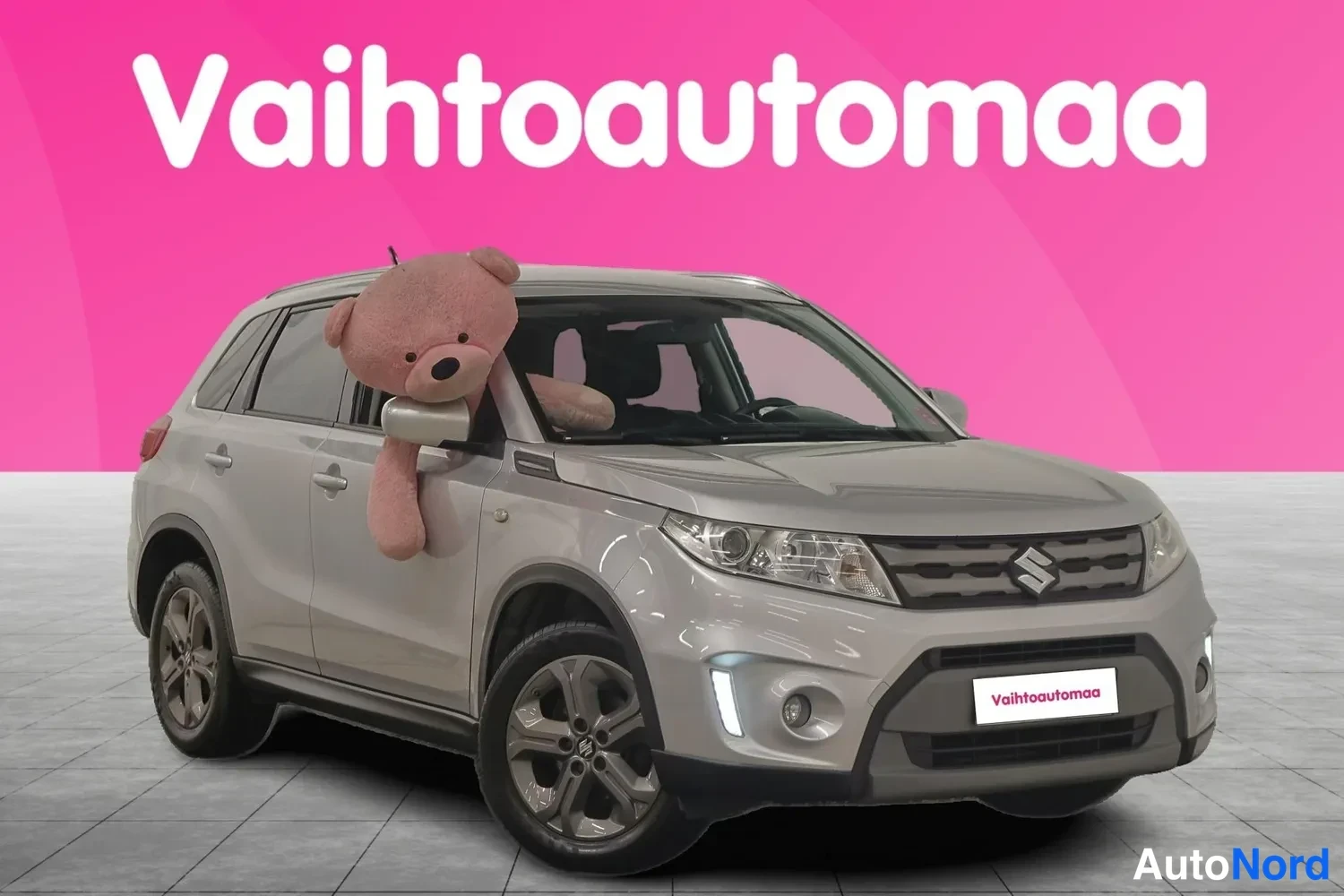 Etuviisto, nokka oikealle: Hopeanharmaa Suzuki etukulmasta oikealta, suuri pehmolelu kiinnitetty matkustajan ikkunaan, tausta vaaleanpunainen Vaihtoautomaa-logolla.