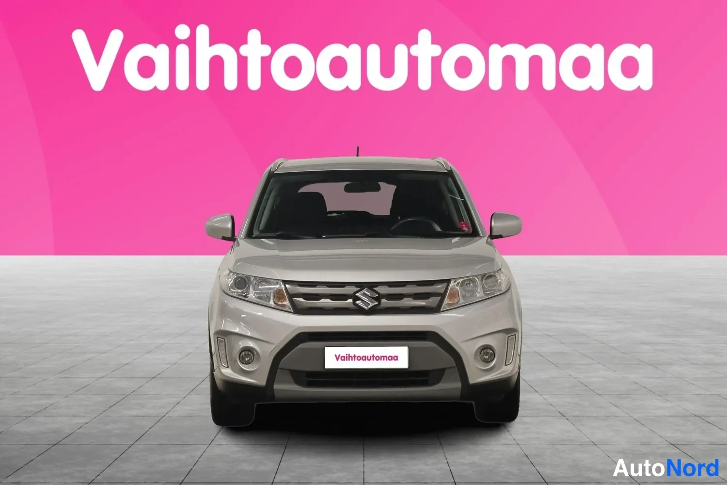 Edestä: Hopeanharmaa Suzuki suoraan edestä, Suzuki-merkki säleikössä, tausta vaaleanpunainen Vaihtoautomaa-logolla.