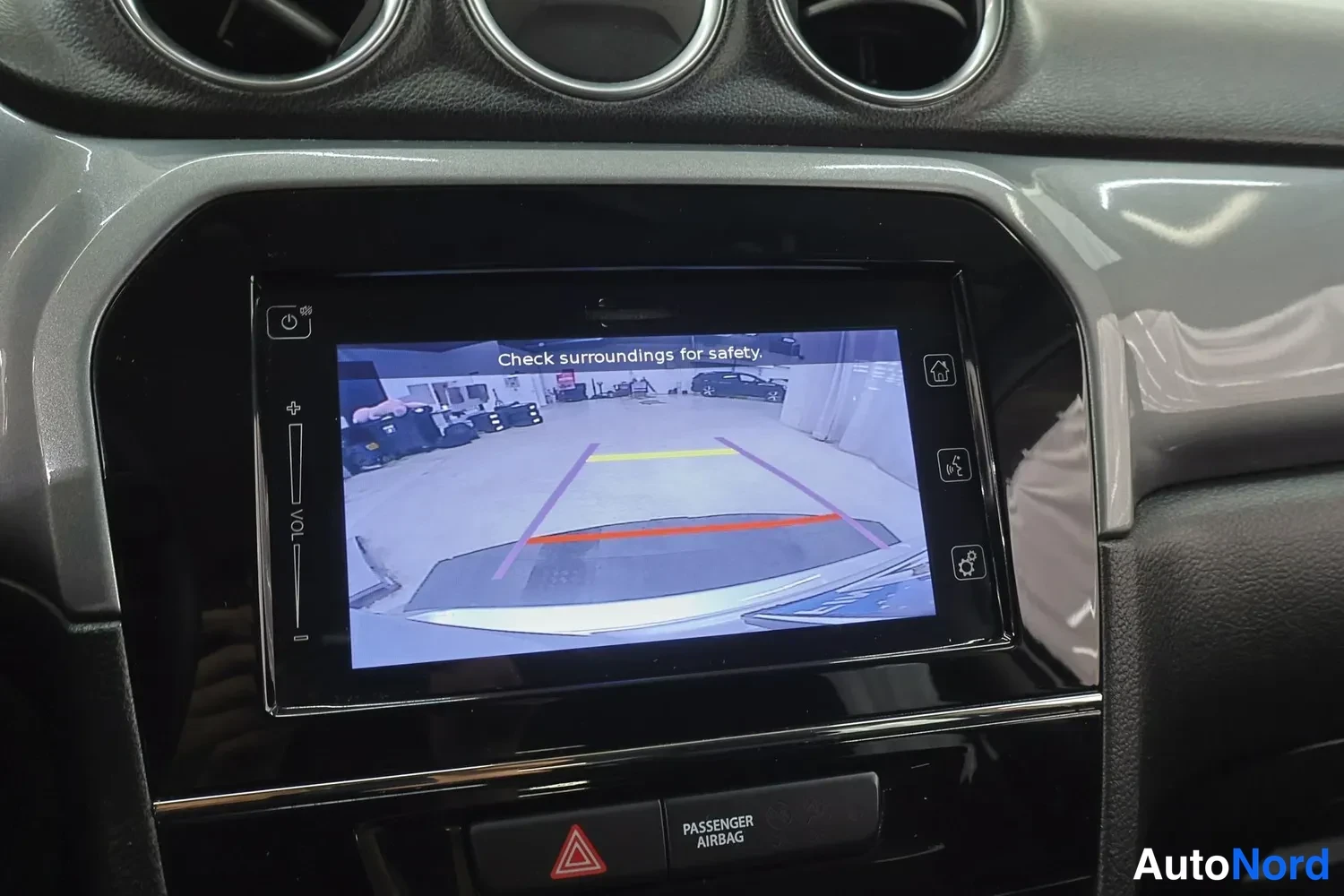 Yksityiskohta: Kojelaudan infotainment-näyttö näyttää peruutuskameran kuvan tekstillä 'Check surroundings for safety.'