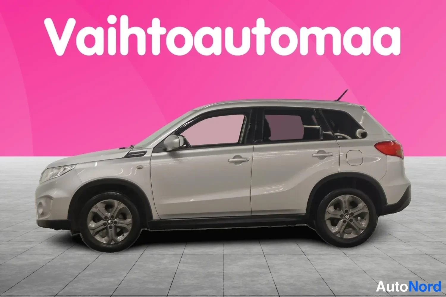 Etuviisto, nokka vasemmalle: Hopeanvärinen Suzuki Vitara sivulta oikealta nähtynä, jokseenkin etuvalo ja kylki täydessä profiilissa pinkkiä studio­taustaa vasten.