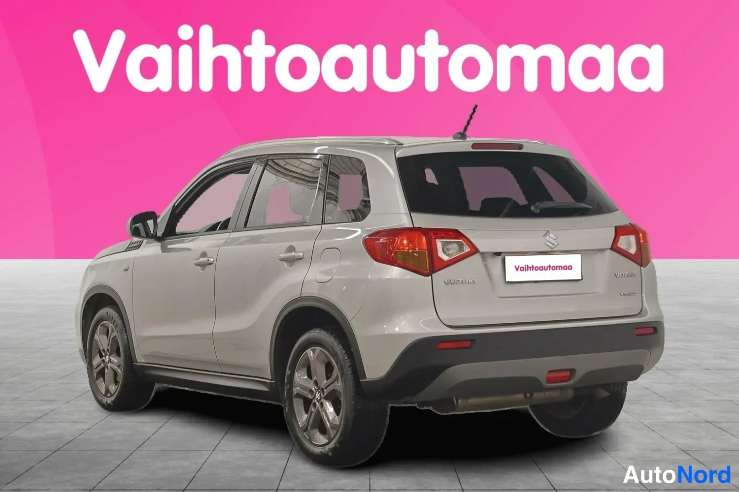 Takaviisto: Hopeanvärinen Suzuki Vitara takakulmalta kuvattuna, taka­valot ja auton vasen kylki näkyvät pinkin taustan edessä.