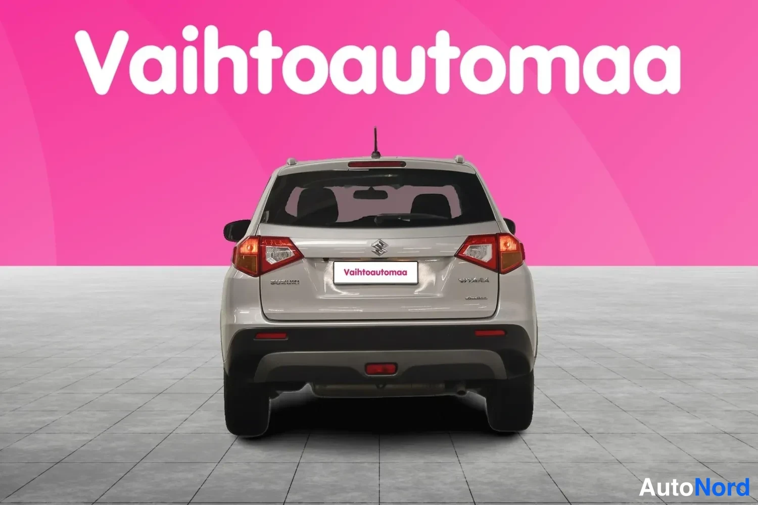 Takaa: Hopeanvärinen Suzuki Vitara suoraan takaa kuvattuna, taka­valot ja takaluukki näkyvät pinkkiä taustaa vasten.