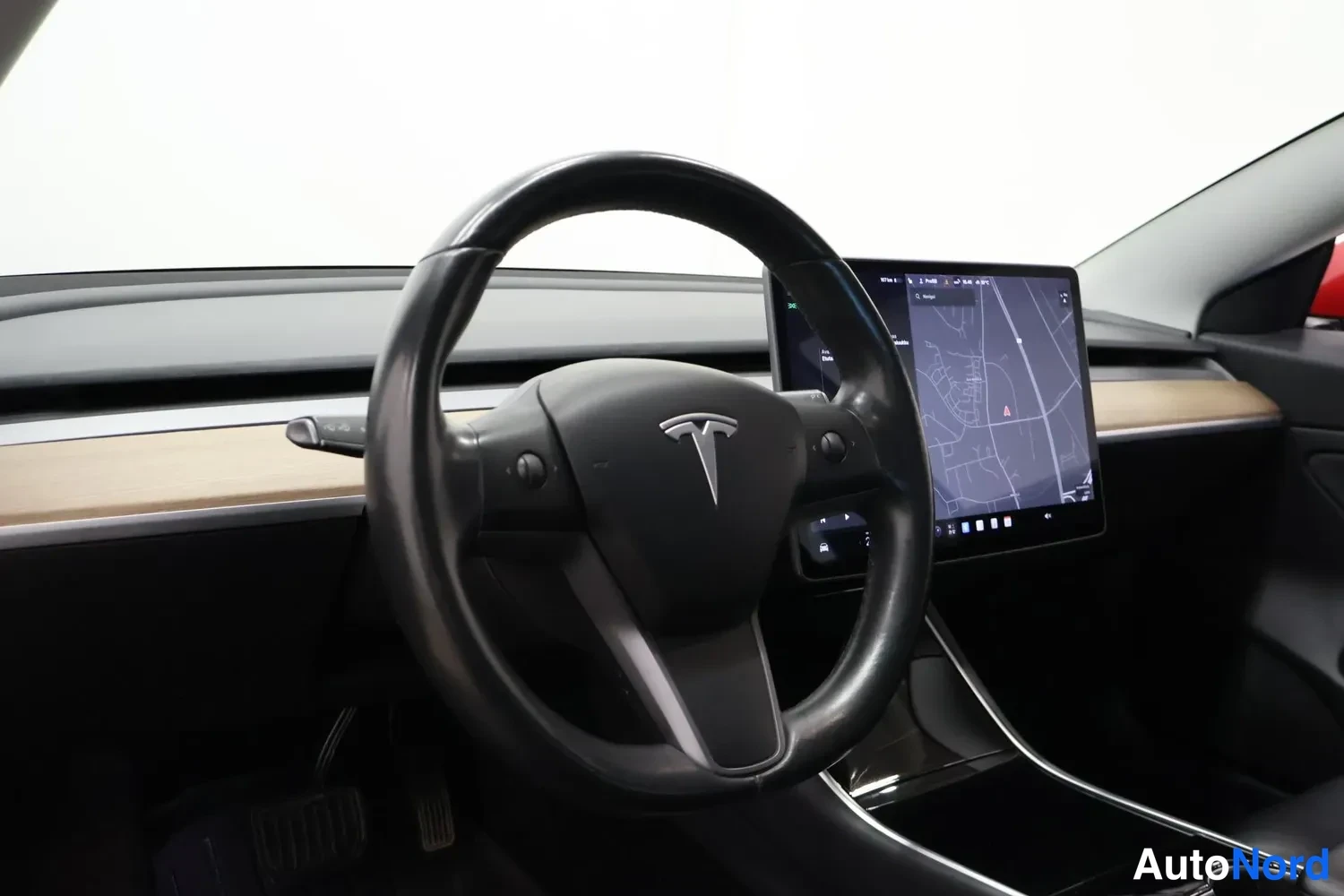 Yksityiskohta: Kuljettajan näkymä: Tesla-logolla varustettu ohjauspyörä, puunvärinen kojelaudan lista ja keskellä suuri karttaa näyttävä kosketusnäyttö.