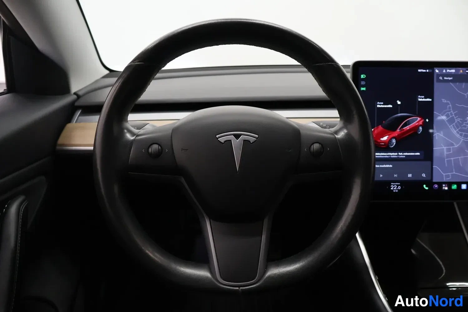 Yksityiskohta: Ohjauspyörän suoraa lähikuvaa Tesla-logolla, oikealla kosketusnäyttö joka näyttää ajoneuvon asetuksia ja karttaa.