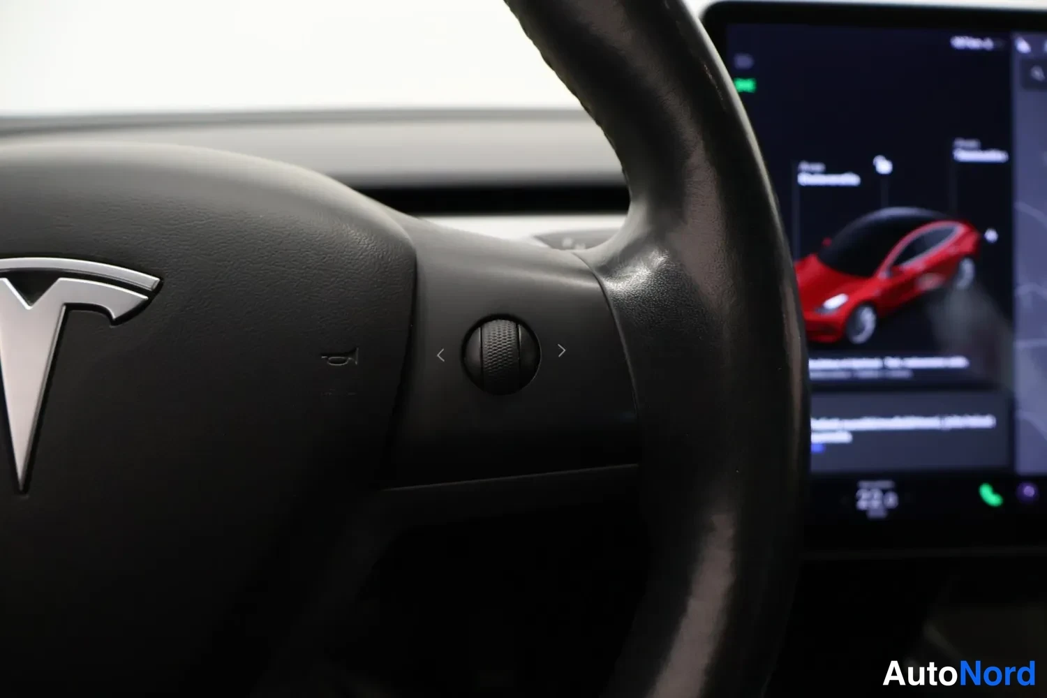 Muu: Lähikuva ohjauspyörän oikeasta kytkinrullasta, taustalla näytöllä punainen Tesla-auto.