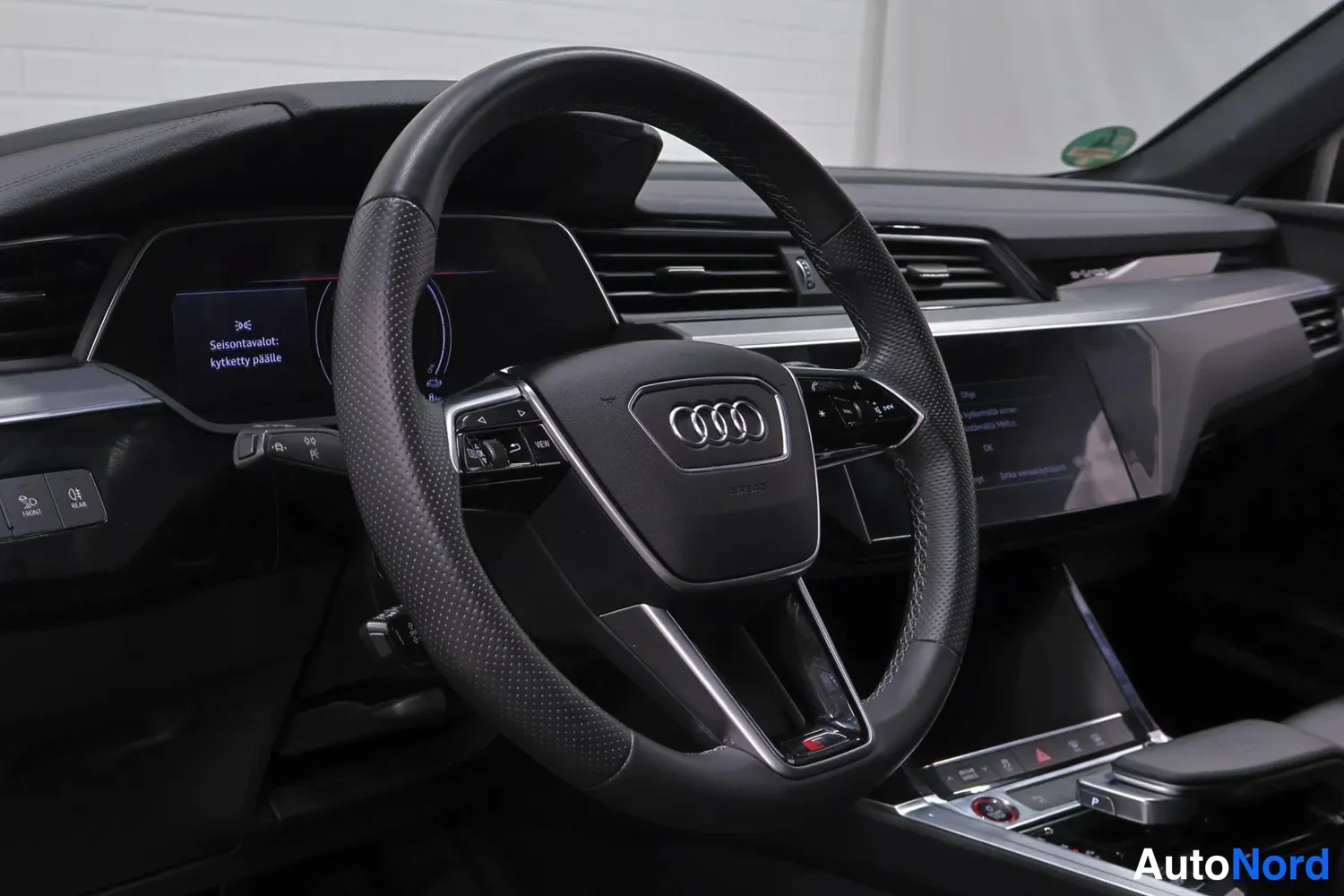 Muu: Audiin ohjaamo: musta nahkainen multifunktio-ohjauspyörä Audi-tunnuksella, kuljettajan digitaalinen mittarinäyttö ja kojelaudan ilmastointisuuttimet.