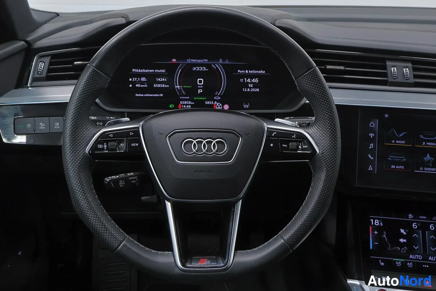 Muu: Ohjaamosta tallikuva: Audi-ohjauspyörä ja mittariston näytöllä lukemat 65858 km, P-vaihde, Radio Metrovol FM, kellonaika 14:46 ja päivä KE 12.8.2026.