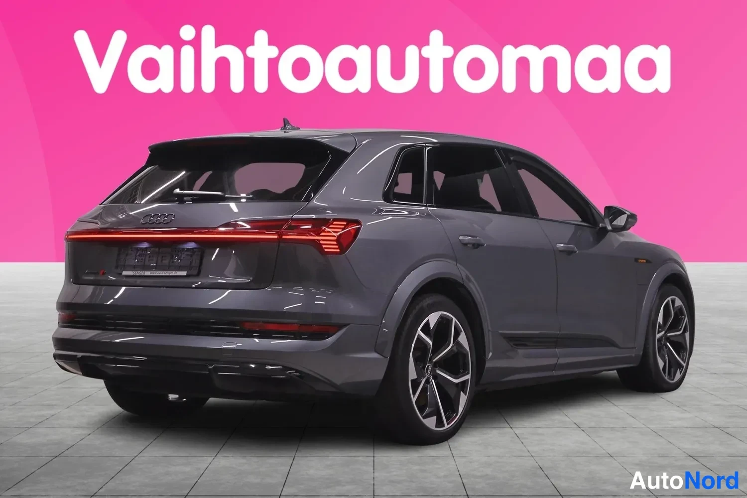 Takaviisto: Tummanharmaan crossoverin takakulmanäkymä: takavalojen yhtenäinen valopalkki, Audi-tunnus takaluukussa ja mustat kevytmetallivanteet, taustalla vaaleanpunainen