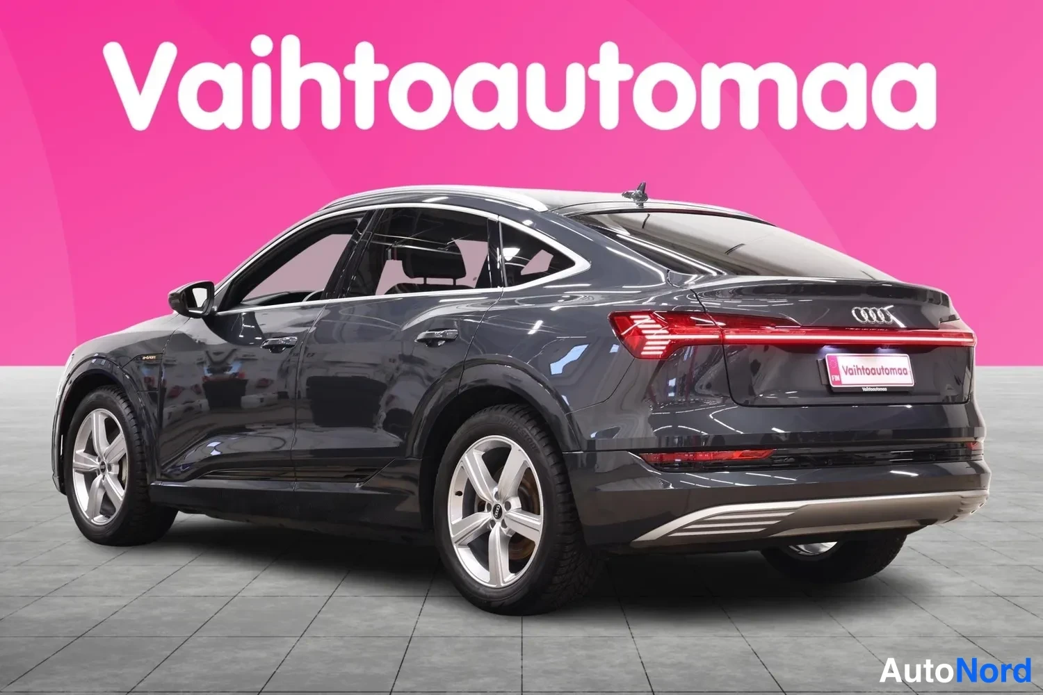 Takaviisto: Audi e-tron Sportback, takakulmanäkymä vasemmalta, tummanharmaa koriväri, LED-takavalot ja vaaleat viisipuolaiset vanteet näkyvissä studiovalossa.
