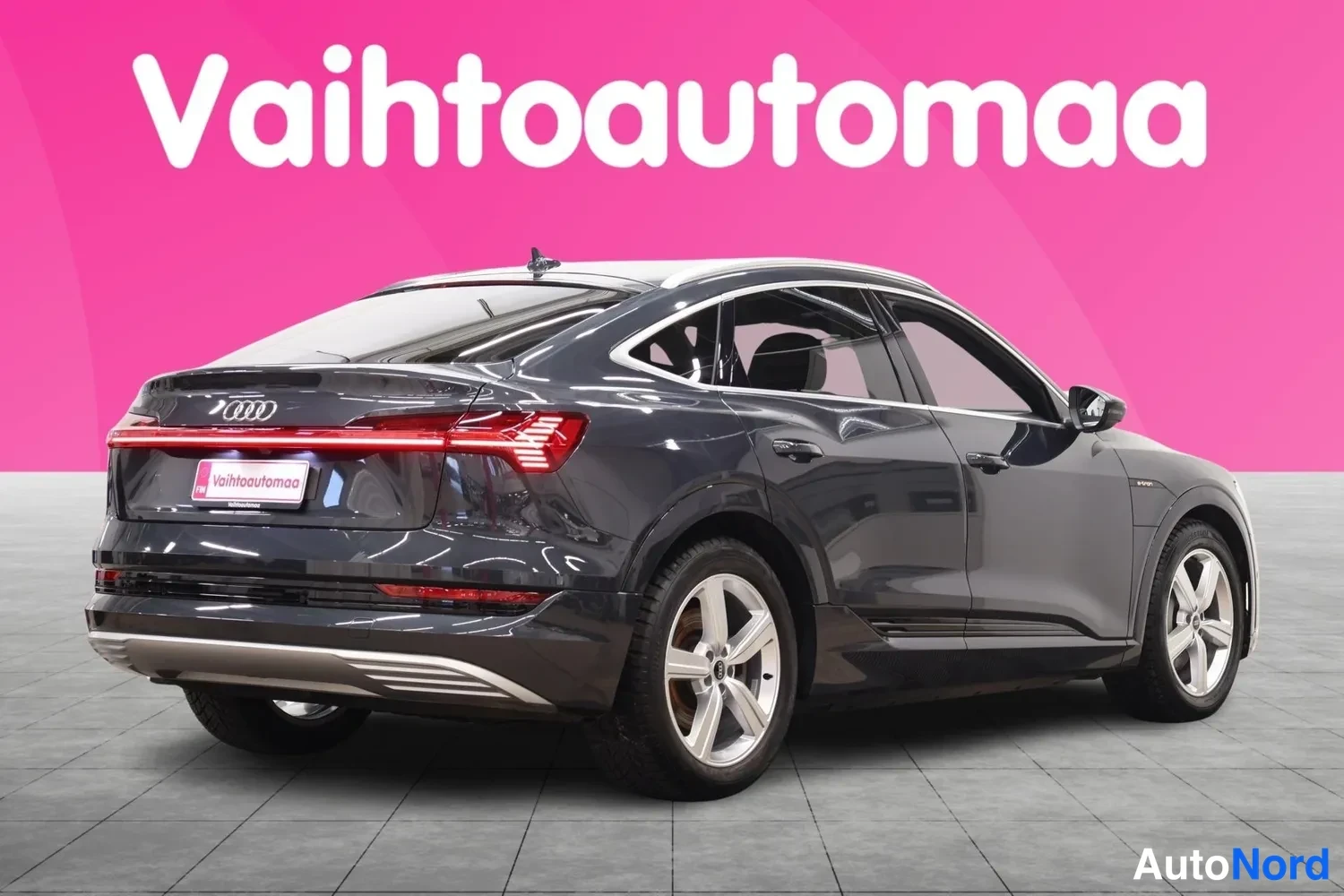 Takaviisto: Audi e-tron Sportback, takakulmanäkymä vasemmalta, tummanharmaa koriväri, takaosan Audi-merkki ja valojuova näkyvissä pinkkiä taustaa vasten.