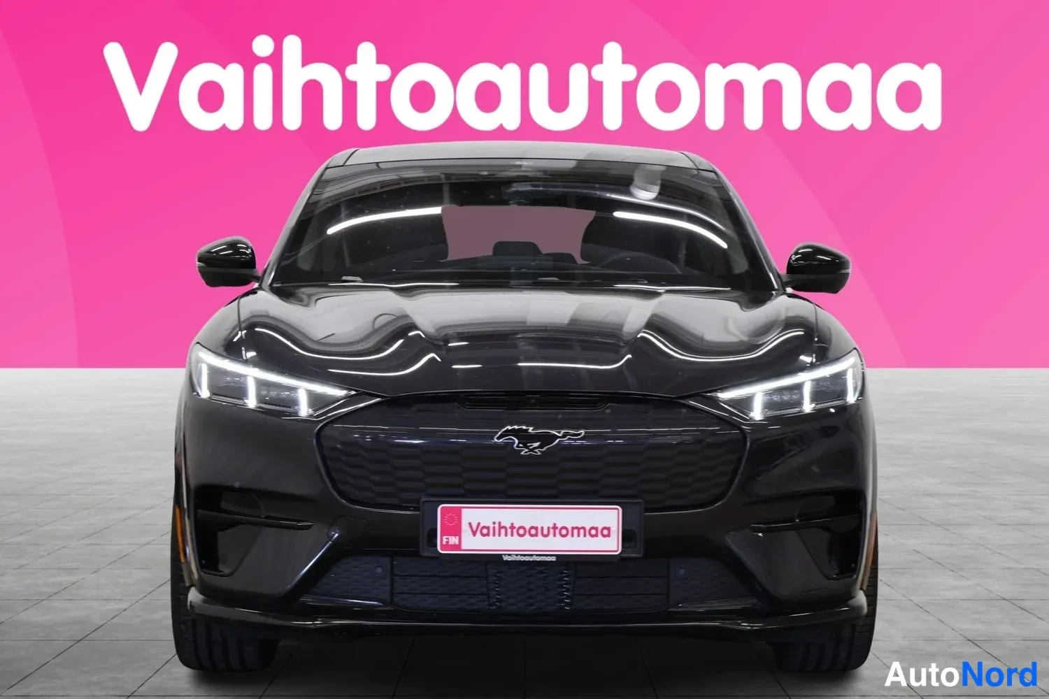 Edestä: Mustan Ford Mustang Mach-E:n suora etukuva, valot päällä ja etumoottorin konepellin alla oleva tavaratila auki.
