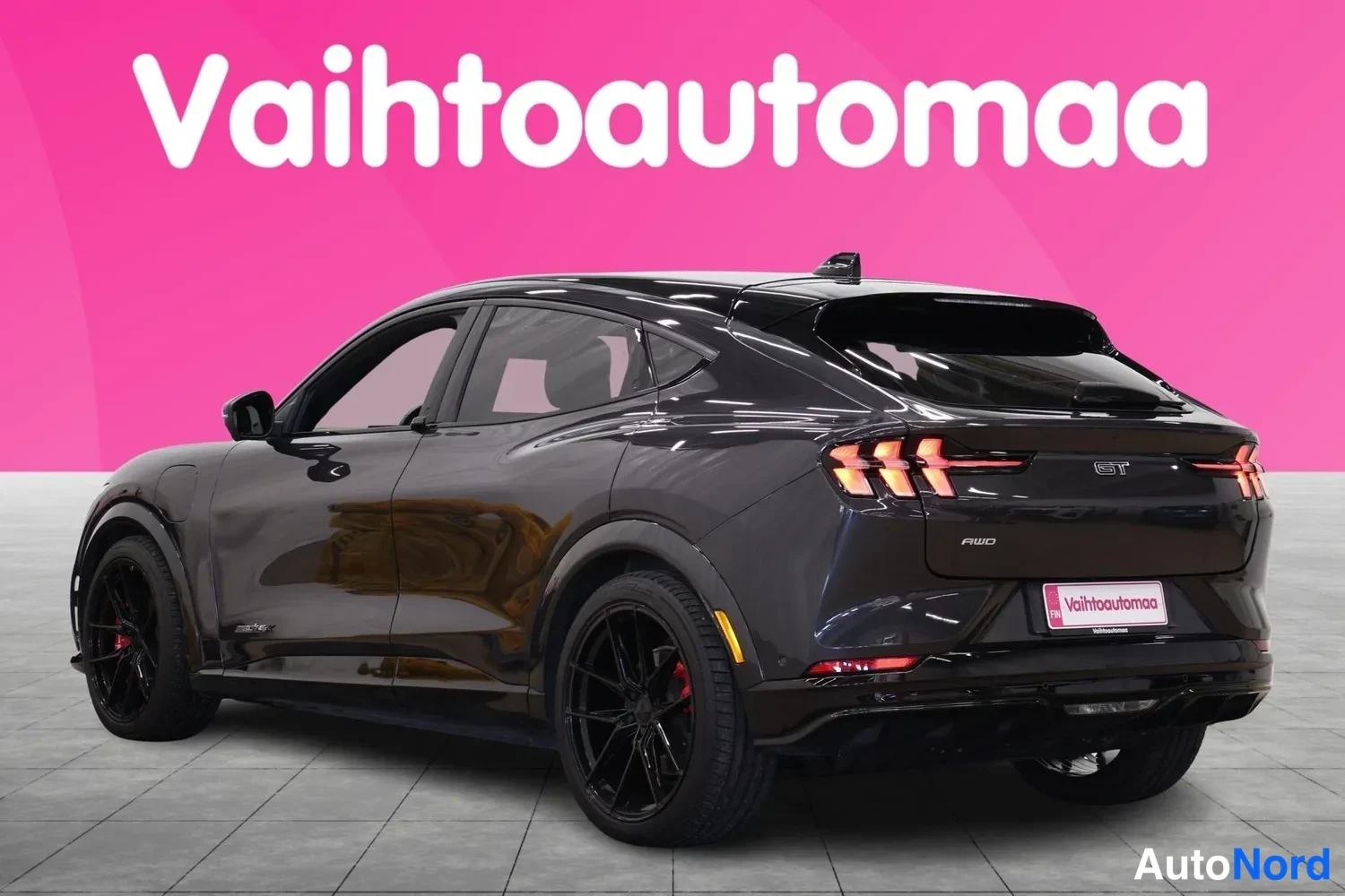 Takaviisto: Mustang Mach-E:n takaosa ja vasen kylki näkyvät vinosta kulmasta mustanharmaana; LED-takavalot ja peräluukun GT-merkki erottuvat.
