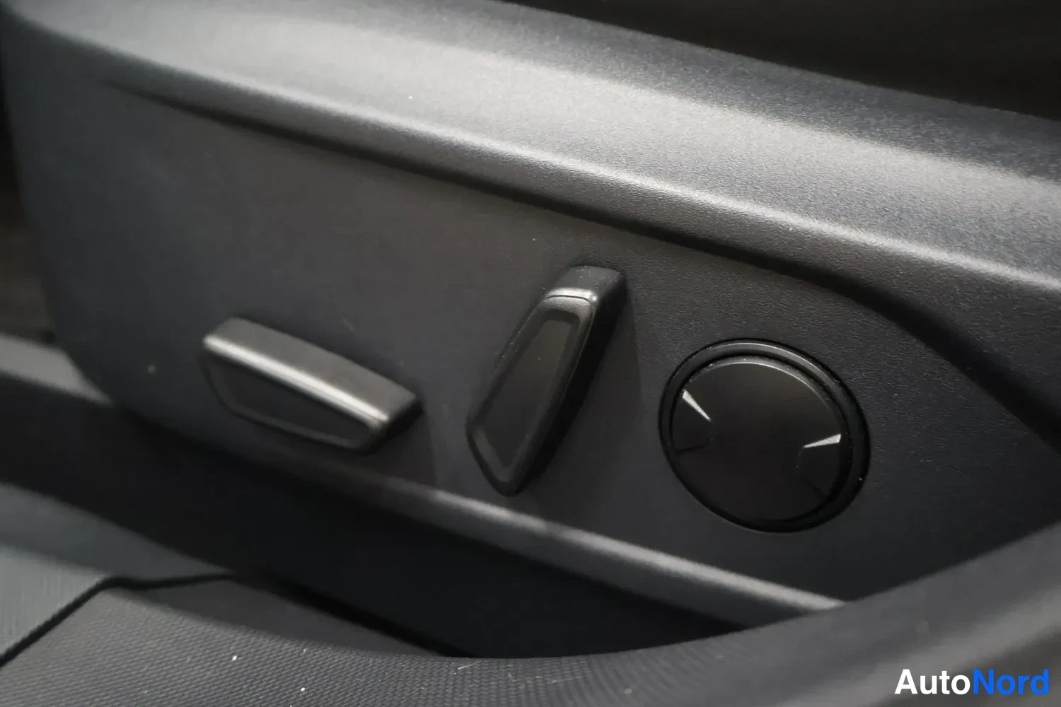 Muu: Sisäkuva: auton oven sivussa olevat istuimen säätönappulat ja pyöreä joystick-tyyppinen säädin, kaikki mustaa muovia.