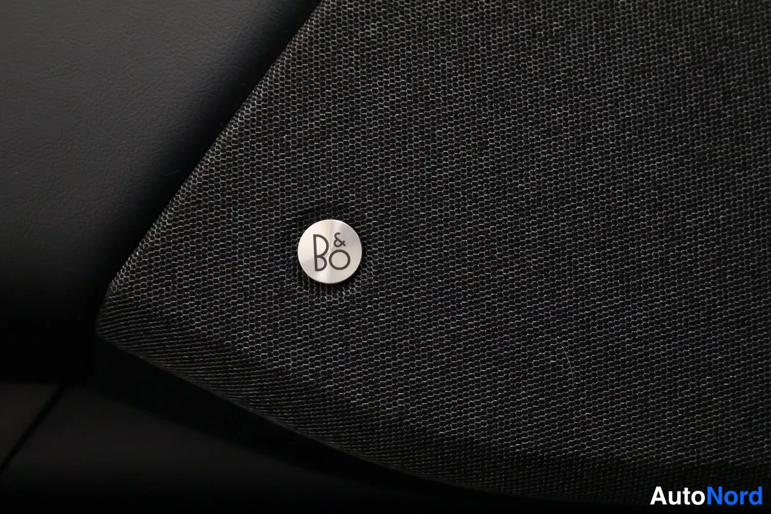Takaa: Kankaalla päällystetty pöytä, jossa kiiltävä B&O-logo — Bang & Olufsen -kaiuttimen läpän verhoilu.