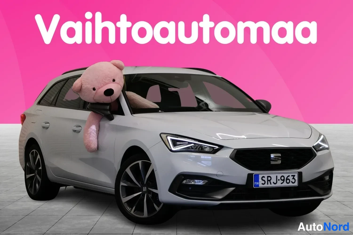 Etuviisto, nokka oikealle: Seat Leon Sportstourer -farmari valkoisena edestä vasemmalta kolmannelta nähtynä, rekisteri, pinkki tausta.
