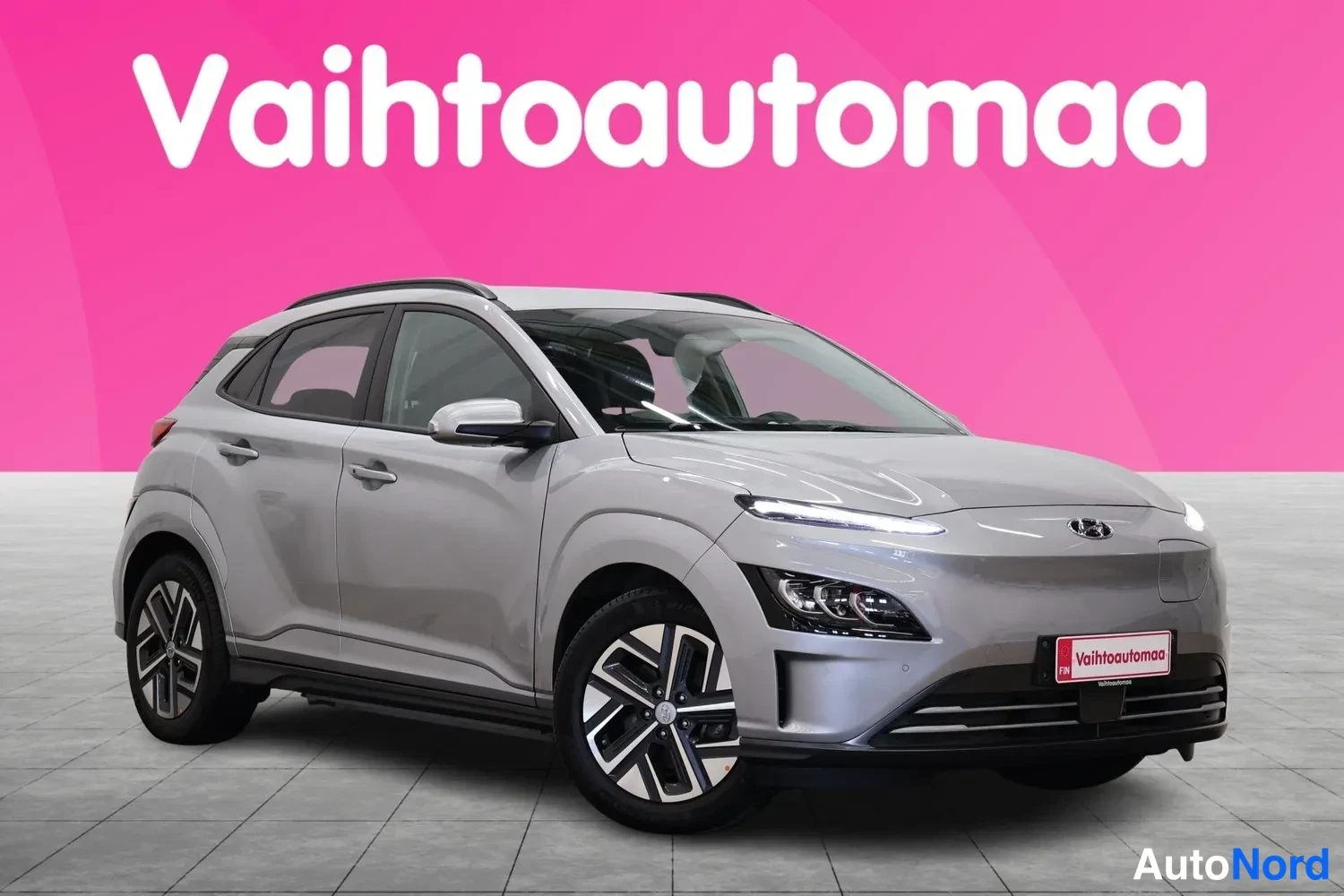 Etuviisto, nokka oikealle: Hyundai Kona Electric -henkilöauto etuviistosta, kevyen harmaa metallinvärinen kori, kylki ja keulanosa näkyvissä pinkissä studioympäristössä.