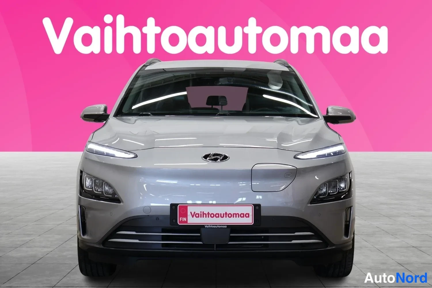 Edestä: Hyundai Kona Electric -henkilöauto suoraan edestäpäin, kevyen harmaa metallinvärinen kori, etuvalot ja etupuskuri näkyvissä.