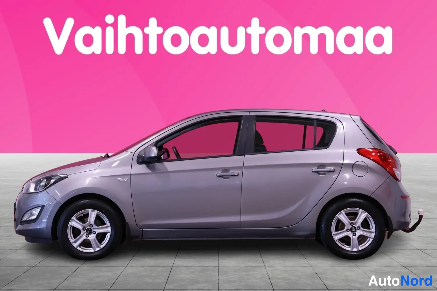Sivulta: Side profile of the silver-grey Hyundai i20 hatchback, showing both wheels, four doors, mirrors, and a rear tow hook attached to a black bumper against a pink b