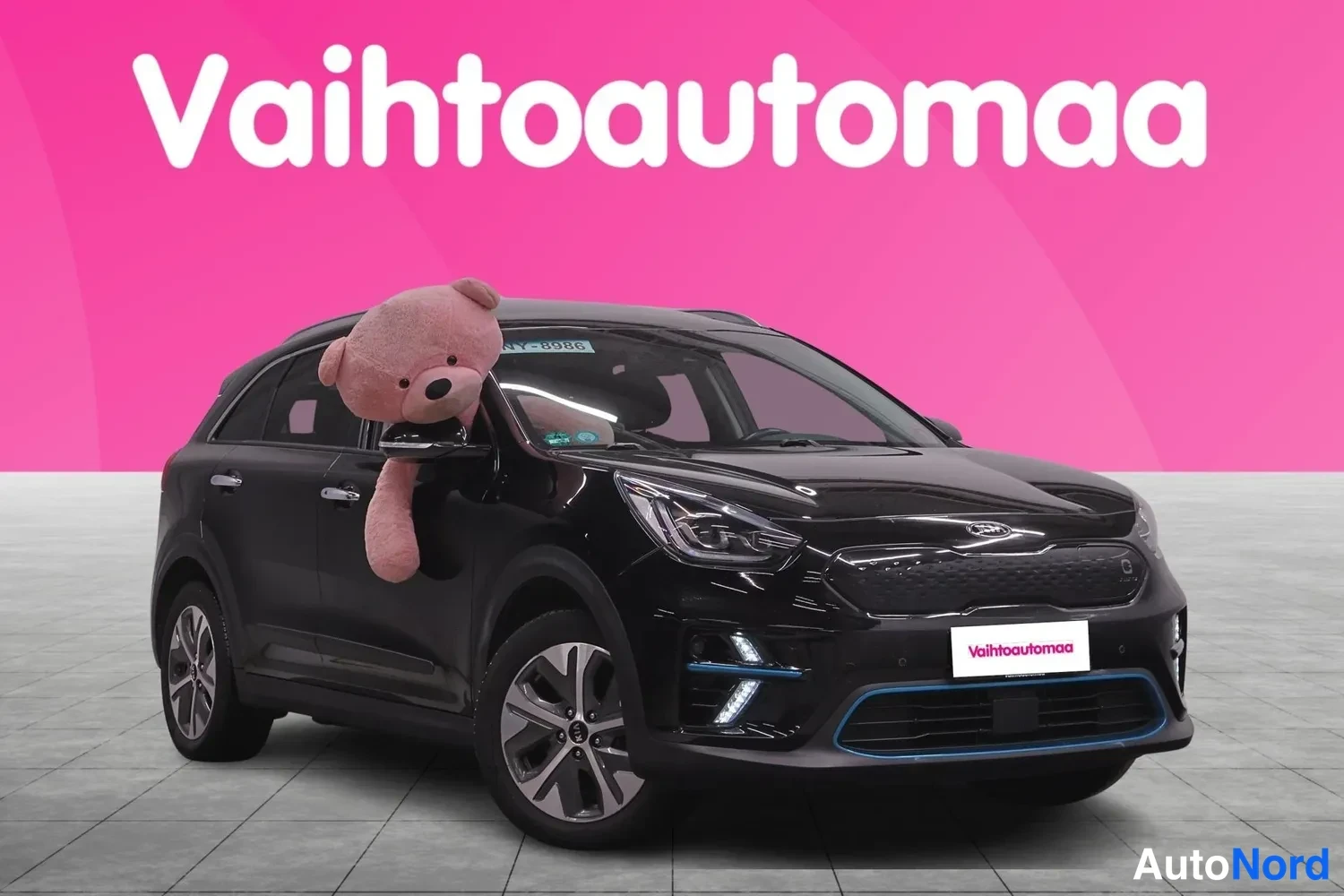 Etuviisto, nokka oikealle: Mustan Kia Niiron etukulmaprofiili oikealta, etusäleikkö ja ajovalot näkyvät, pinkki tausta.