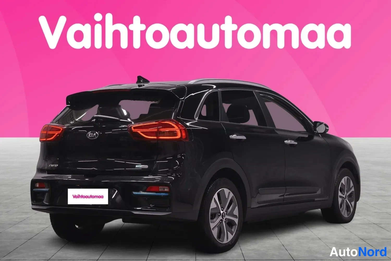 Takaviisto: Musta Kia Niro takaa kolmanteen neljään. Auto seisoo vaalealla laattoine lattiassa pinkin taustan edessä. Takavalot ovat punaiset ja puskurin alapuolella on sin