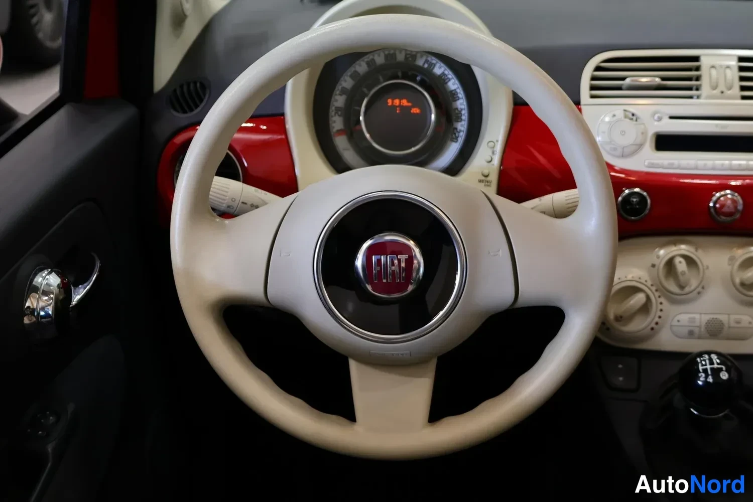 Muu: Autojen sisätilakuva: beige ohjauspyörä Fiat-tunnuksella, takana mittaristo, punainen korostuspaneeli kojelaudassa, ilmastoinnin säätönupit ja manuaalivaihteen