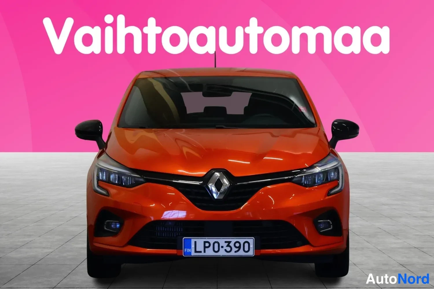 Edestä: Oranssi Renault Clio suoraan edestä päin, Renault-merkki ja -rekisterikilpi näkyvissä, vaaleanpunainen tausta.