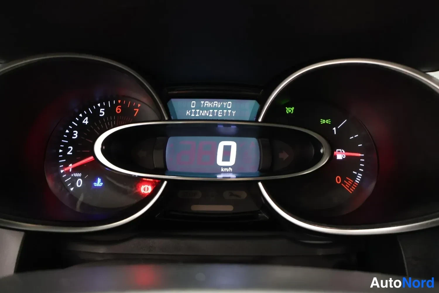 Yksityiskohta: Koeajon mittaristo: analoginen kierroslukumittari ja polttoainemittari, keskellä digitaalinäyttö 0 km/h ja viesti 'D TAKAVYO KIINNITETTY'.