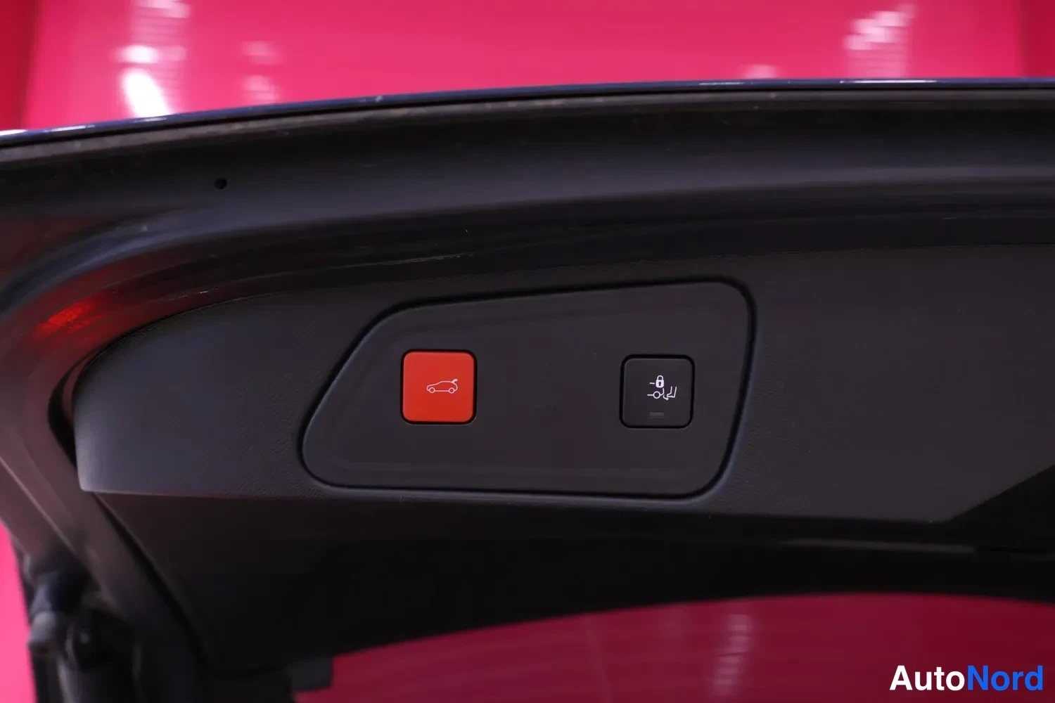 Yksityiskohta: Interior rear view of open boot lid showing two buttons on black plastic lining: red button with open trunk icon and adjacent button with car-on-jacks symbol in