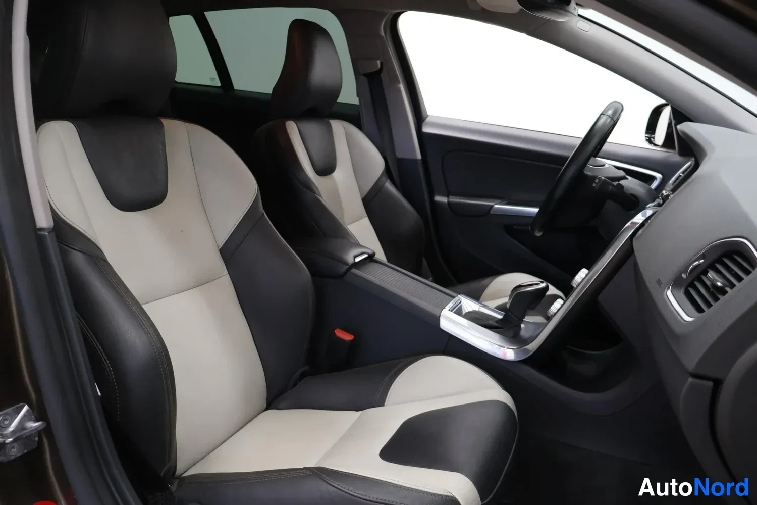 Muu: auton etupenkki ja keskikonsoli musta-beige-riskiupholsterilla, ohjauspyörä ja vaihdekeppi näkyvissä.