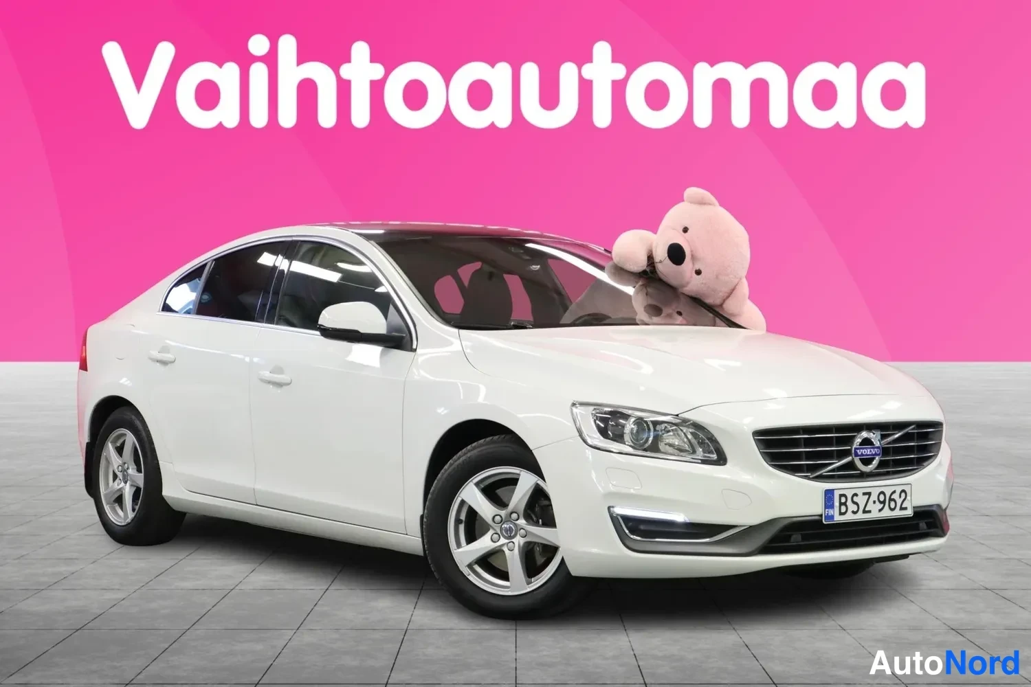 Etuviisto, nokka oikealle: Valkoinen henkilöauto vinosti etualalla, etusäleikkö ja rekisterikilpi näkyvät, taustalla pinkki mainosseinämä.