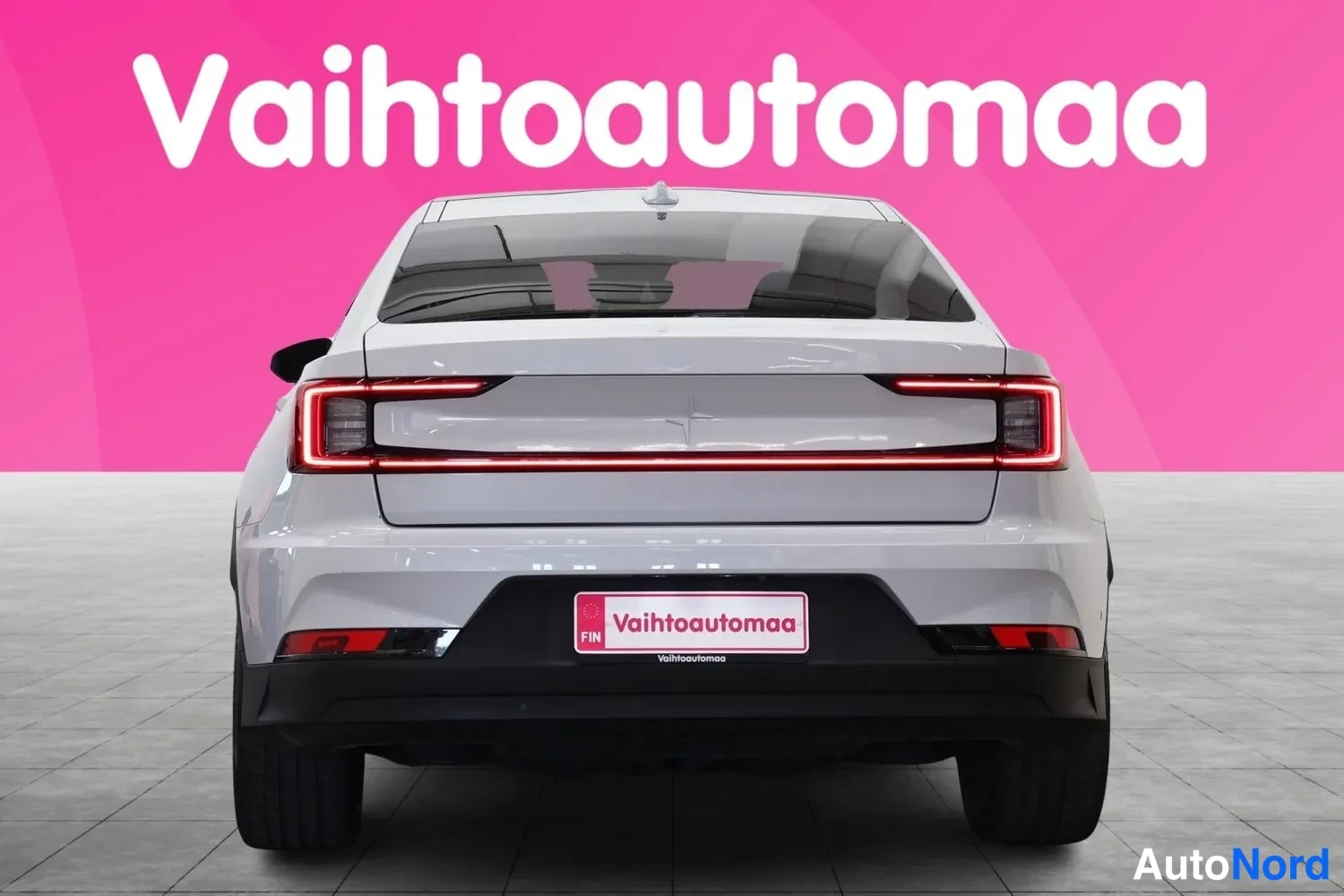 Takaa: Auton takaosa suoraan takaa: punaiset takavalot, valkoinen kori ja rekisterikilven paikalla Vahtoautomaa-kyltti, taustalla pinkki mainosseinämä.