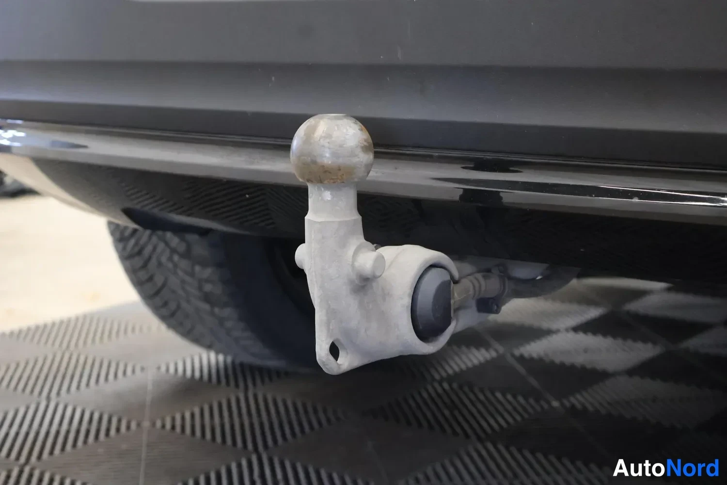 Yksityiskohta: Takaympyrän ja vetokoukun lähikuva. Vetokoukun pallo on irrotettu ja koukku on kiinnitetty takapuskurin alapuolella olevaan kiilaan. Koko laite on harmaa metall