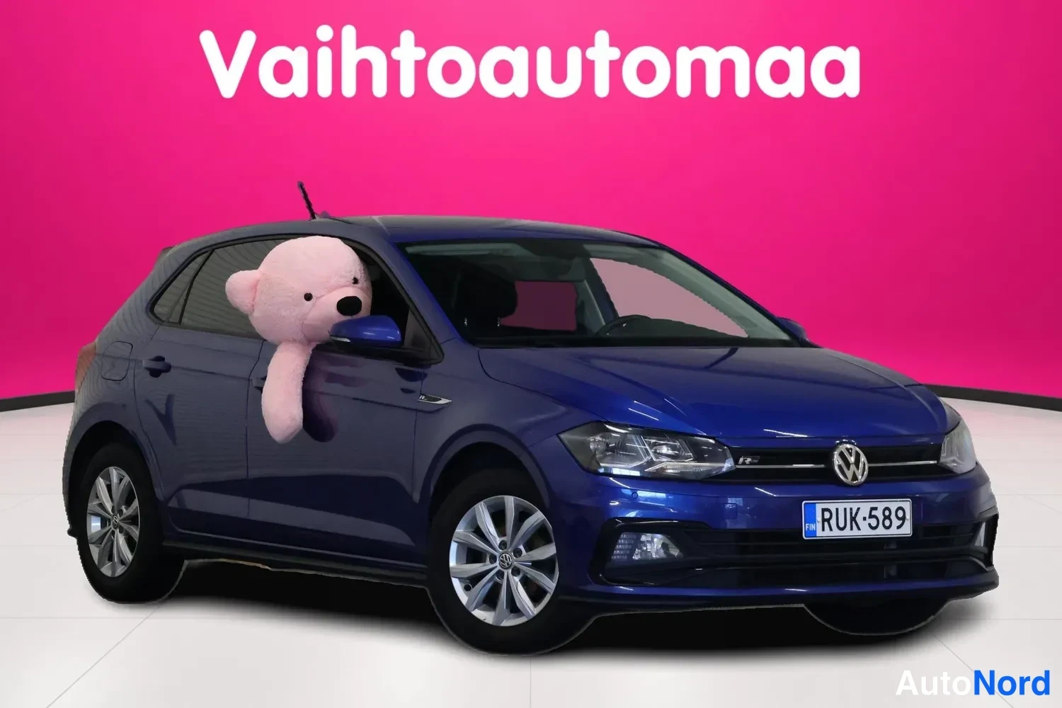 Etuviisto, nokka oikealle: Sininen Volkswagen Poli etukulmasta oikealta, rekisterimerkki, teksasarja taustalla.