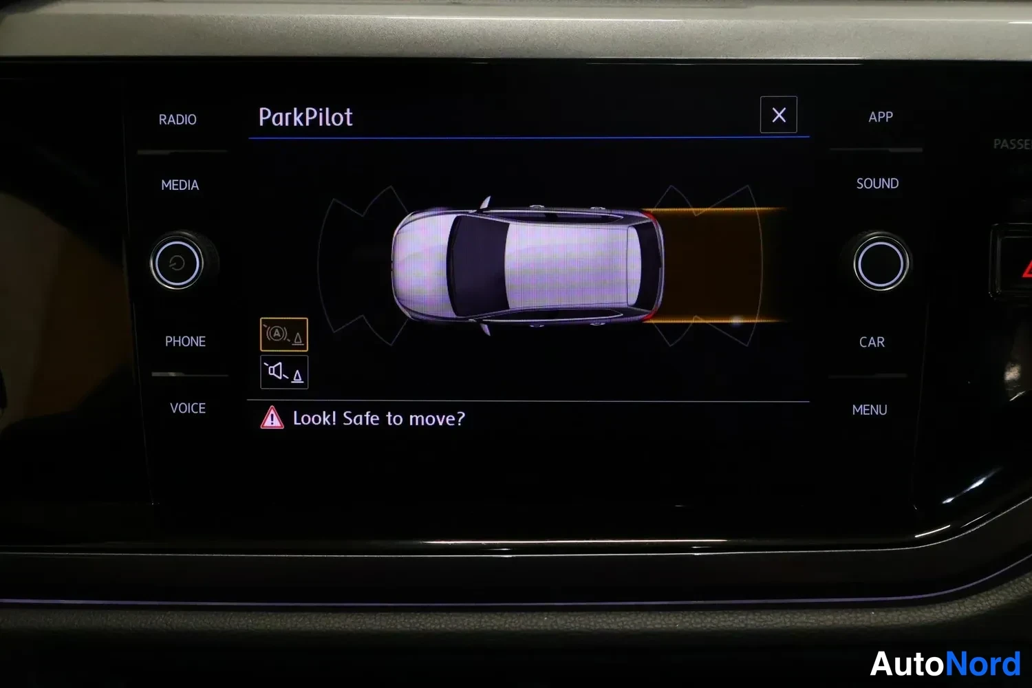 Muu: Volkswagenin keskikonsolin ruutu näyttää ParkPilot-säätövalvonnan graafin, jossa autoa ja takaisia antureita on merkitty, sekä varoituksen 'Look! Safe to move?'