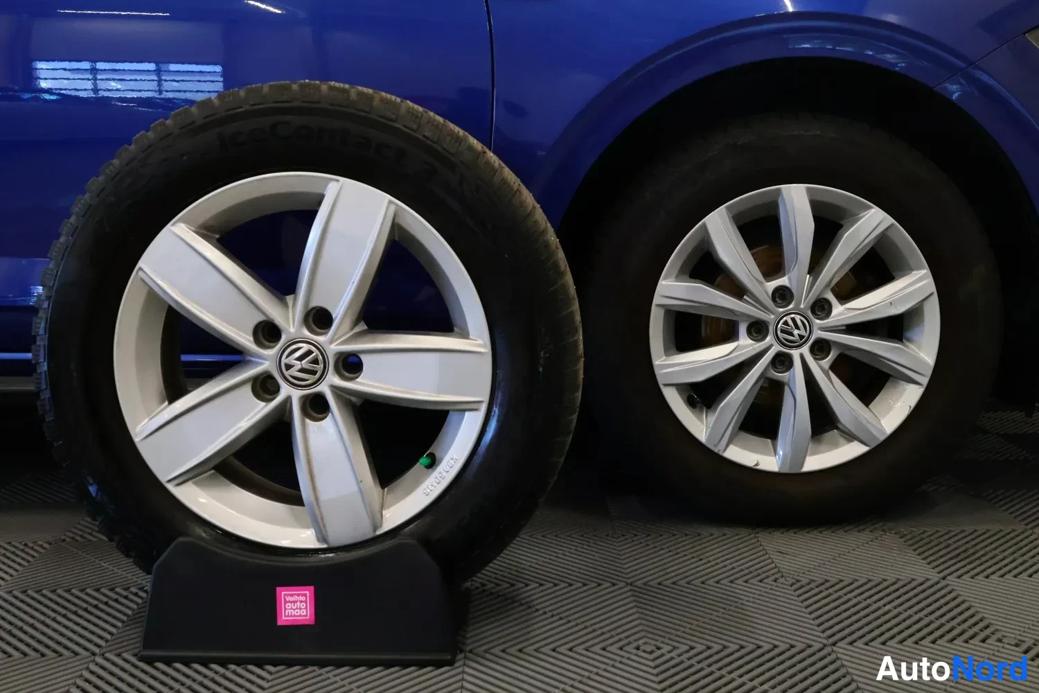 Sivulta: Sinisen auton kylki ja takapyörä Volkswagen-tunnuksella; edessä telineessä seisova varapyörä hopeisella vanteella ja IceContact-renkaalla.