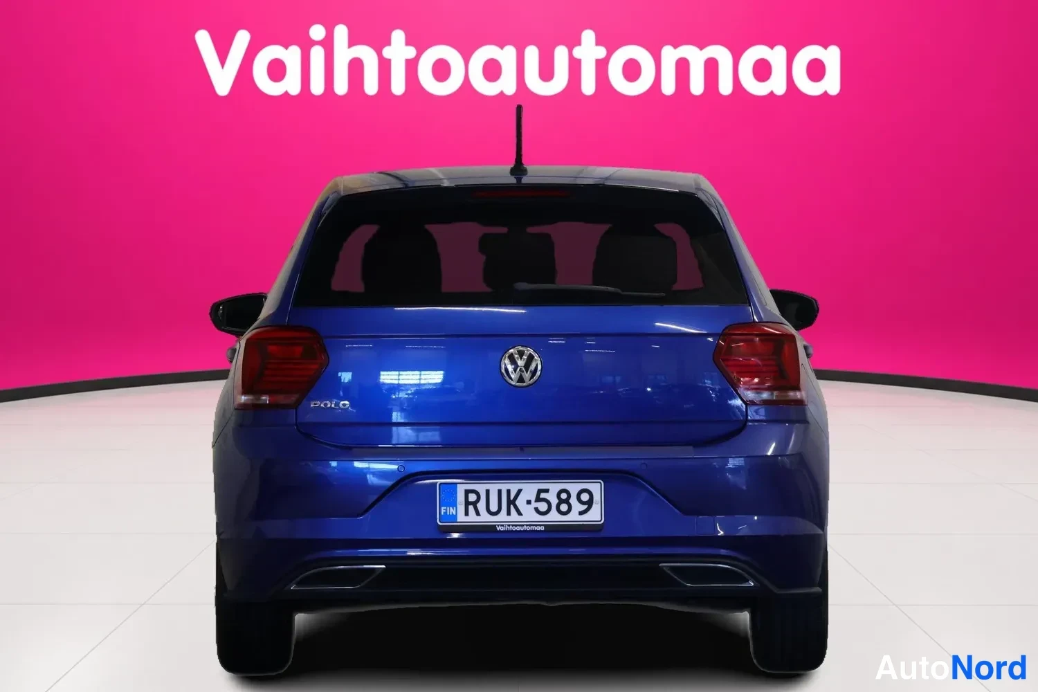 Takaa: Sininen Volkswagen Polo suoraan takaa, rekisterikilpi, vaaleanpunainen tausta.