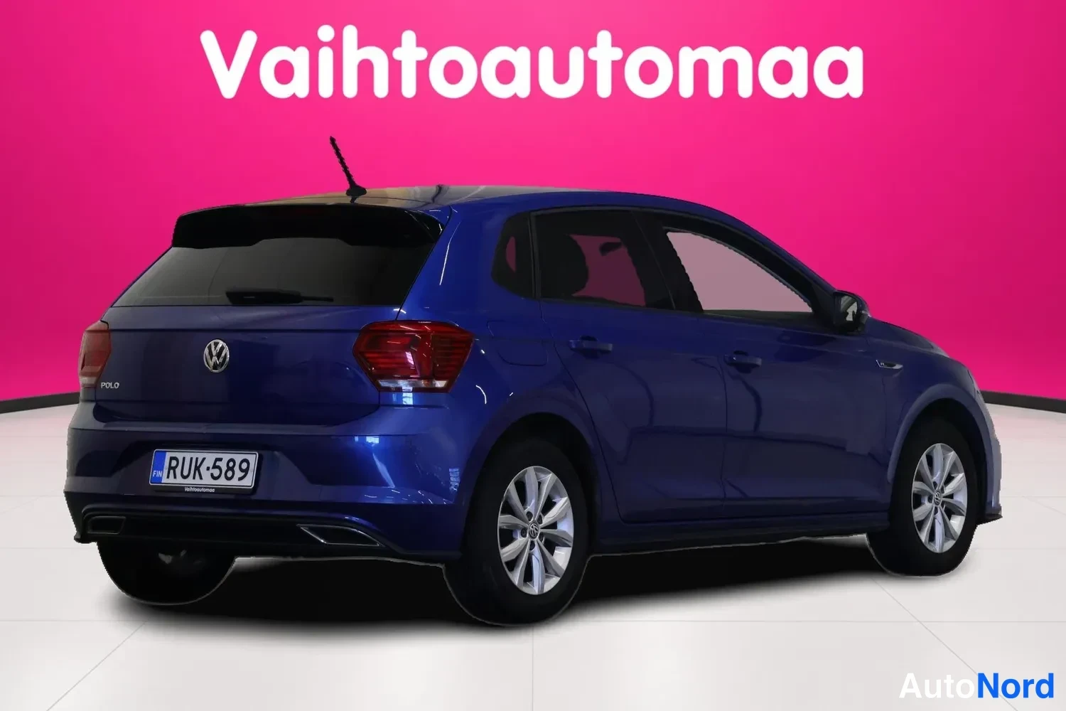 Takaviisto: Sininen Volkswagen Polo takaviistosta oikealta, rekisterikilpi, vaaleanpunainen tausta.