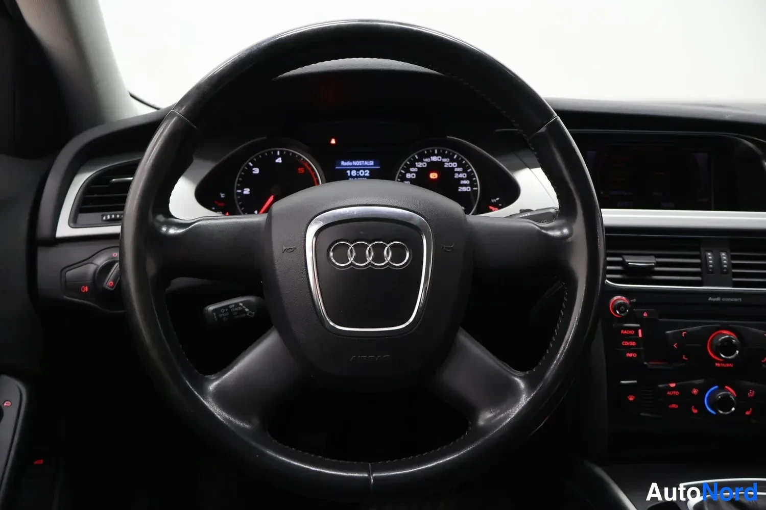 Muu: Suoraan ohjauspyörään kohdistuva kuva: Audi-merkki keskellä, mittaristo palaa, laitteen näytössä teksti 'Radio MOSAIK' sekä kello 16:02.