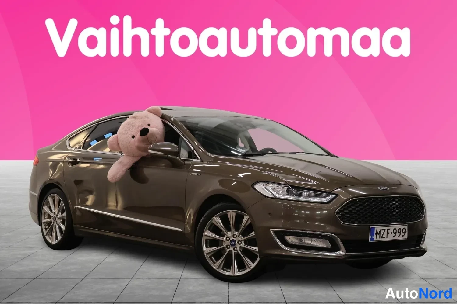 Etuviisto, nokka oikealle: Ruskea Ford Mondeo -henkinen sedan etuvastakulmasta oikealta, rekisteritunnus, suuri pehmolelu ikkunassa, pinkki tausta.