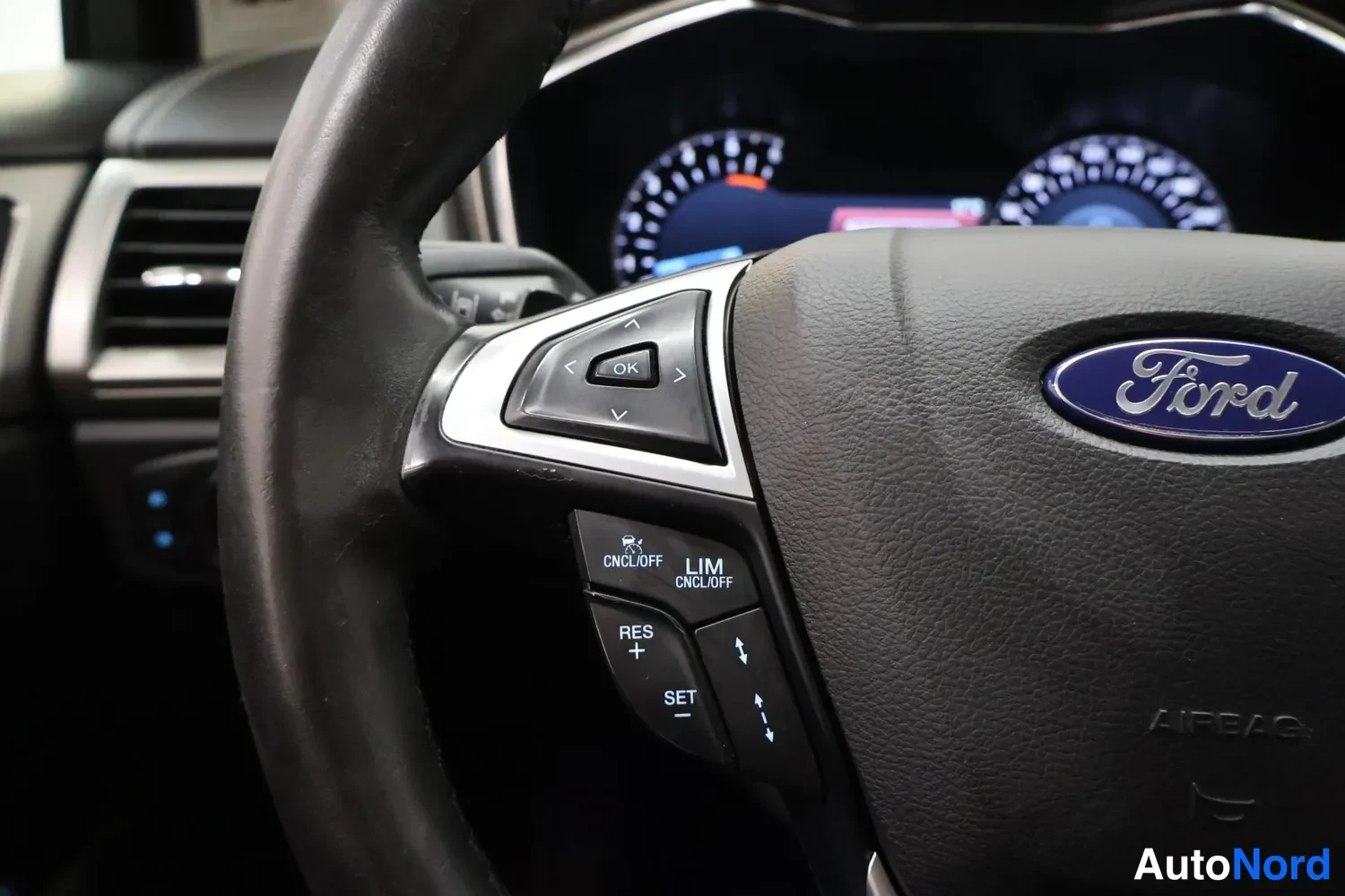 Muu: Lähikuva Ford-ohjauspyörästä ja sen vasemmasta painikeplokista, joissa matkustajan turvatyynyn AIRBAG-merkki ja nopeudensäädön napit.