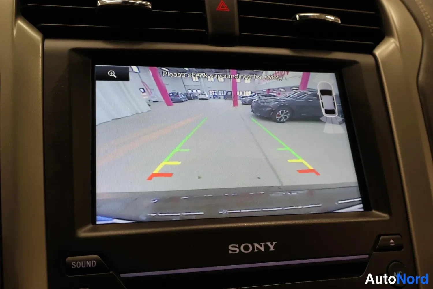 Muu: Auton kojelaudan Sony-merkkisen infotainment-näytön lähikuva. Näytöllä on peruutuskameran kuva hallitilasta, vihreästä, keltaisesta ja punaisesta pysäköintiviiv