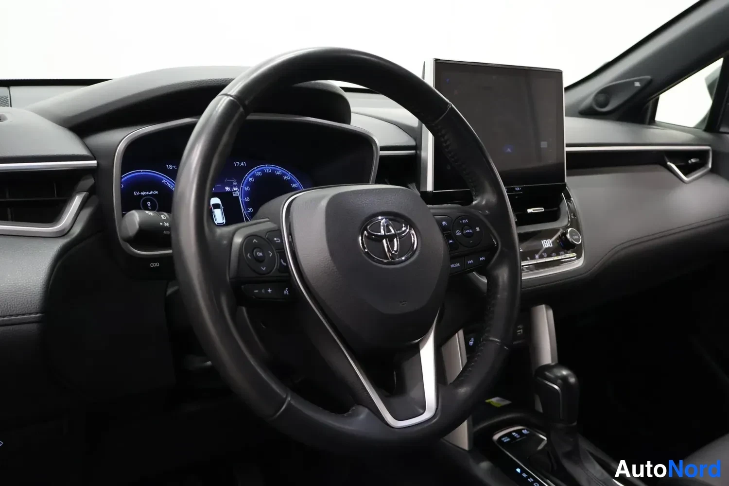 Yksityiskohta: Kuljettajan näkymä ohjaamoon: Toyota-merkillä varustettu ohjauspyörä, osittain sähköinen mittaristo ja keskikonsolin infotainment-näyttö.