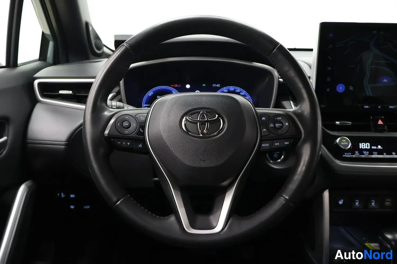 Yksityiskohta: Ohjauspyörä ja kojelauta suoraan edestä: Toyota-logo, monitoiminapit, digitaalinen mittaristo ja kartta näytöllä.