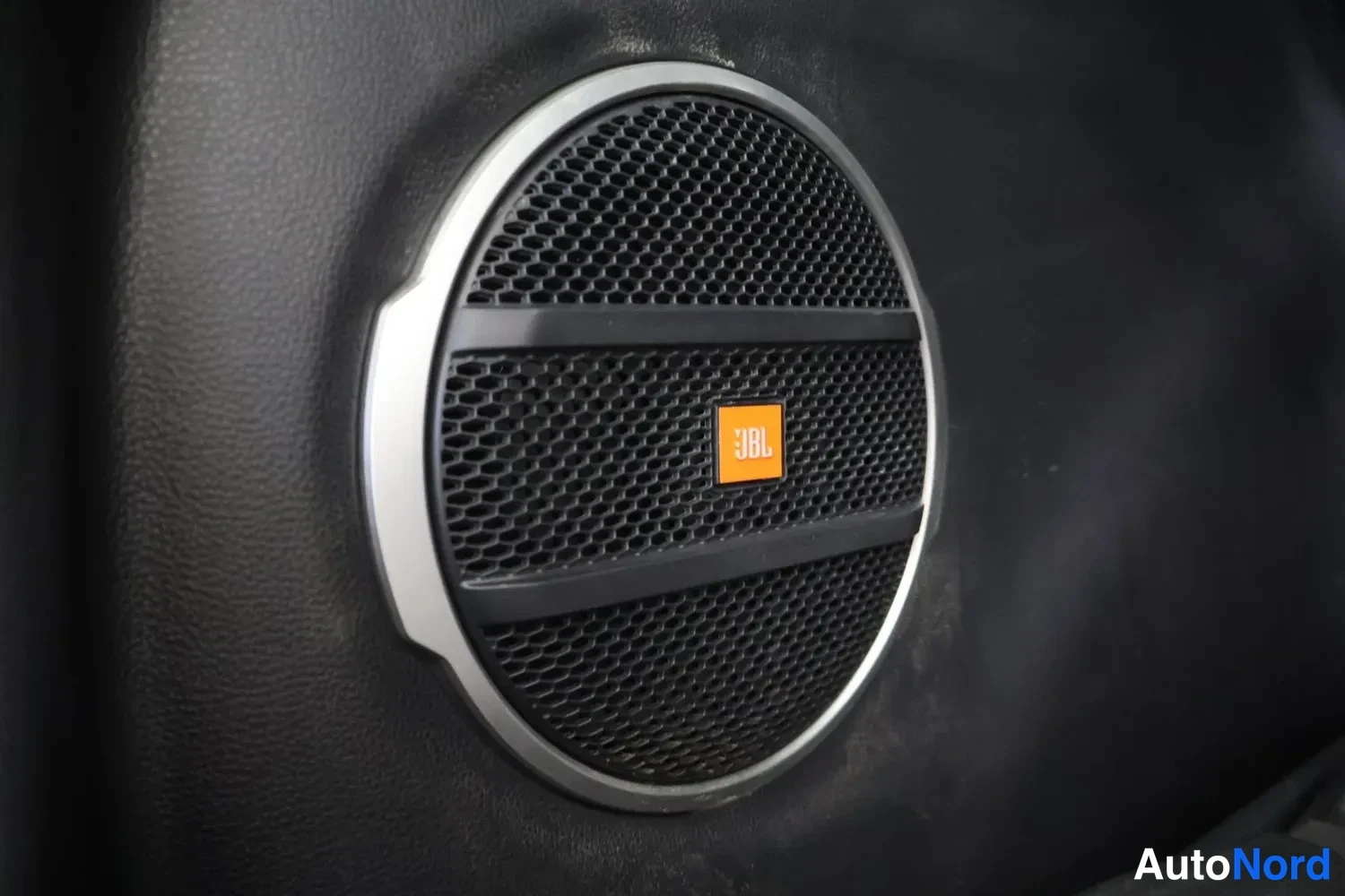 Yksityiskohta: Sisätilan lähikuva: ovipaneeliin asennettu pyöreä JBL-kaiutin, jossa hopeanvärinen kehys ja oranssi JBL-logo mustassa ritilässä.