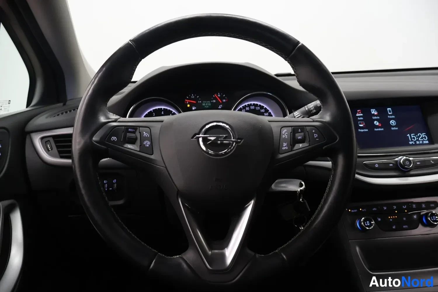 Muu: Suora kuva ohjauspyörästä: Opel-tunnuksellinen ohjauspyörä, mittarit syttyneenä ja keskinäyttö näkyy oikealla; kyljessä on jotain tekstiä.