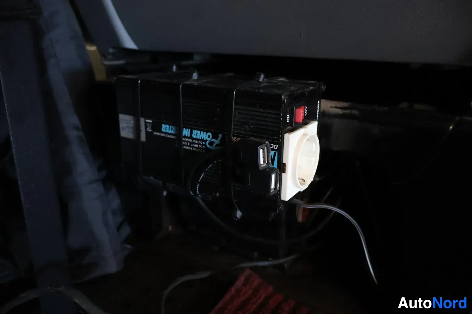 Muu: Musta invertteri/teholähde, jossa punainen kytkin, saksalainen pistorasia ja USB-liittimet, kiinnitettynä auton lattialle.