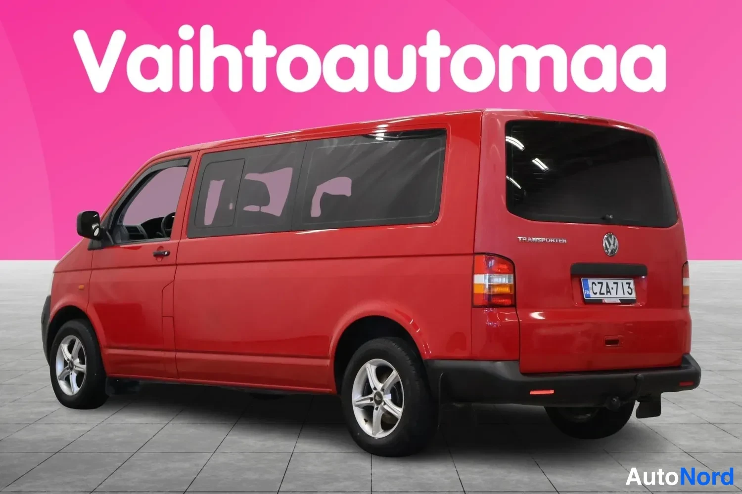 Takaviisto: Punainen Volkswagen Transporter -pakettiauto takaviistosta vasemmalta, rekisterikilpi, pinkki tausta.